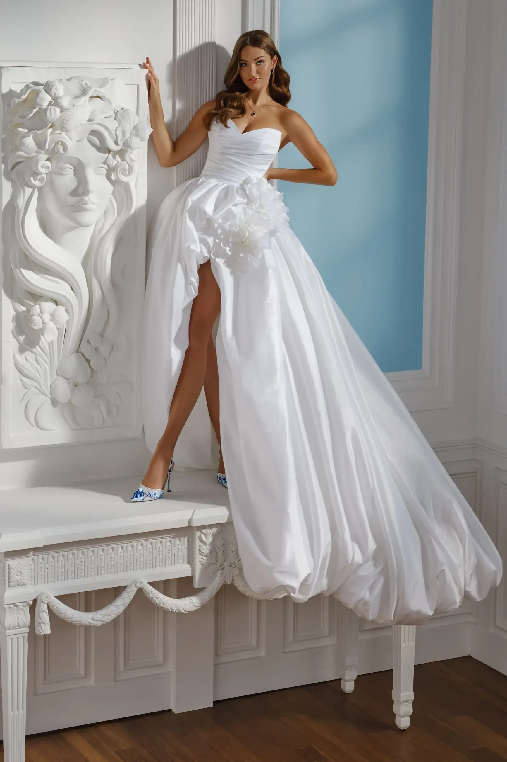 A woman in a flowing white gown stands on a white ornate table, with one leg forward. The background features light blue walls and a floral relief.