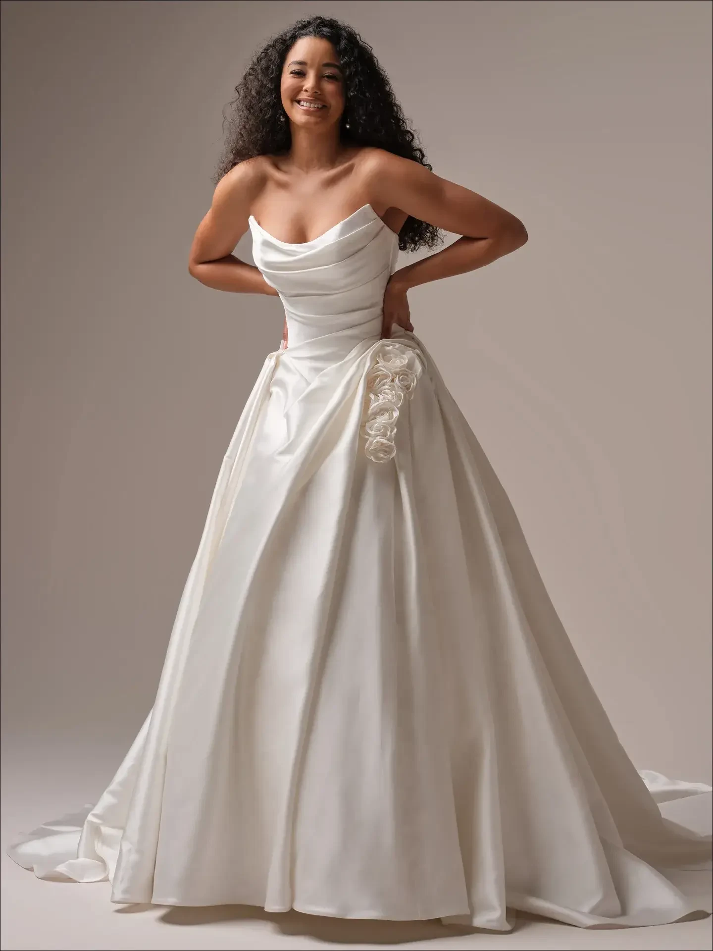 A woman wearing a white wedding dress with a strapless design and a floral detail at the waist, posing elegantly against a neutral background.