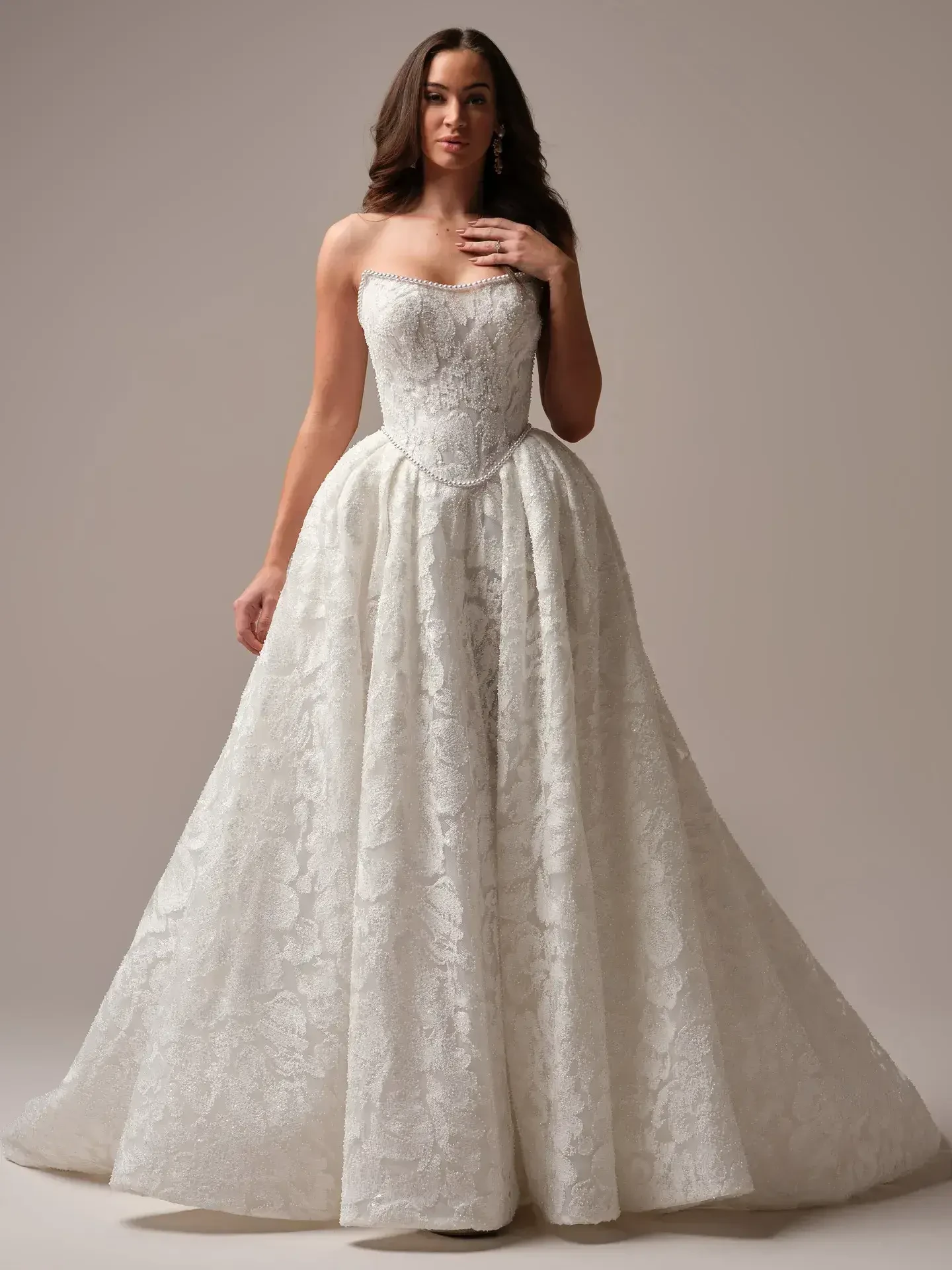 A woman wearing a strapless, floor-length wedding dress with intricate lace detailing. The gown features a fitted bodice and a voluminous skirt.