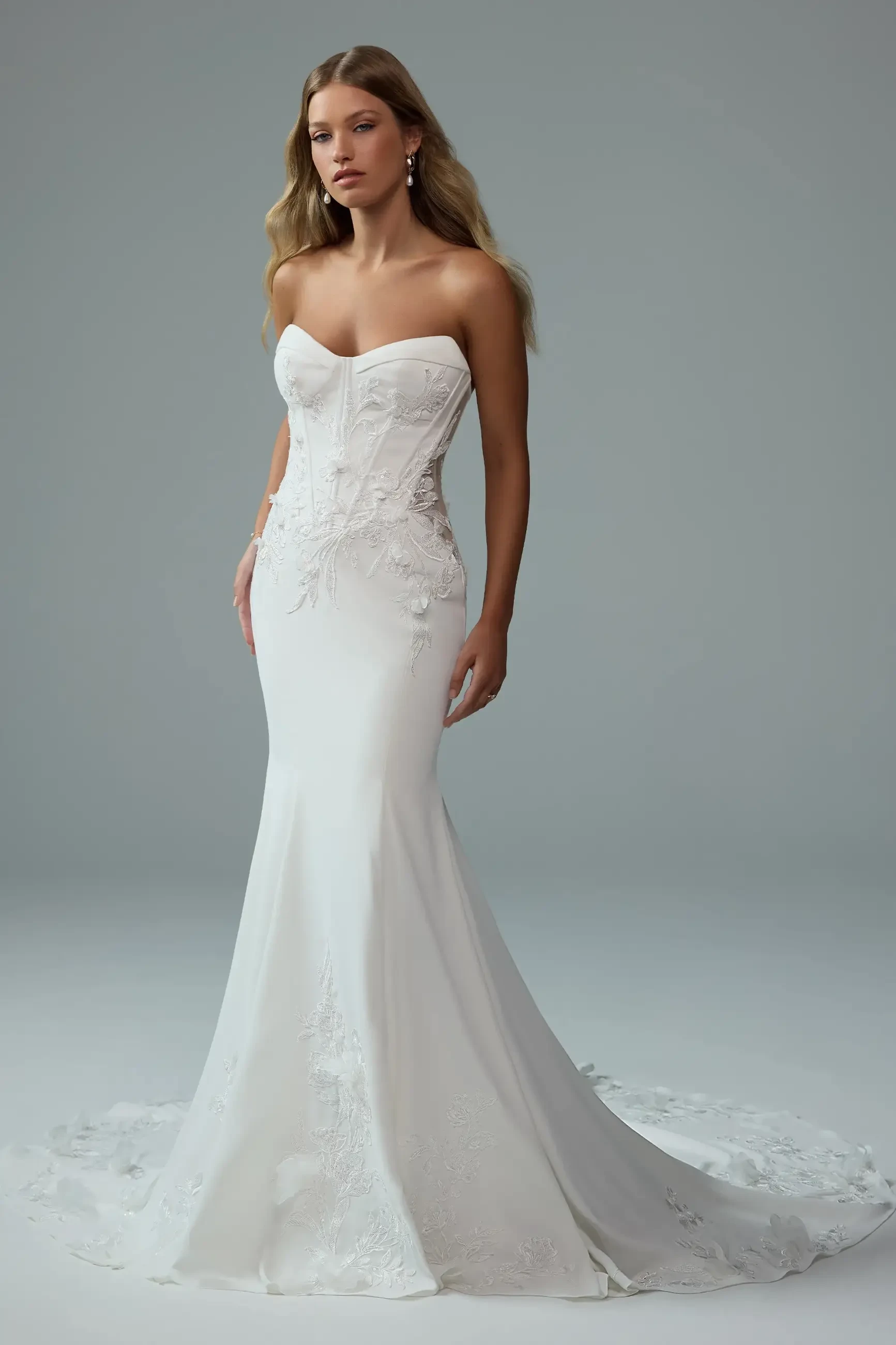 A model wearing a strapless white wedding dress with floral embellishments, standing against a neutral background.